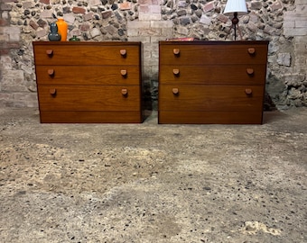 May include: Two matching wooden dressers with four drawers each. The dressers are a warm brown color with rounded wooden drawer pulls. The background is a brick and stone wall and a concrete floor.