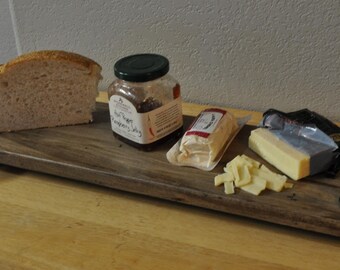 May include: A wooden serving board with bread, a jar of preserves, cheese, and cheese cubes. The board has metal handles and a tag attached. The items are arranged on a wooden surface.