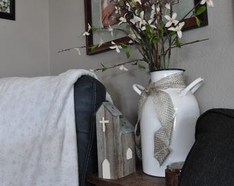 May include: A white enamel milk jug vase with a silver ribbon, holding white flowers and greenery. A small wooden church with a cross sits on a dark wooden table. A framed photo hangs on the gray wall.
