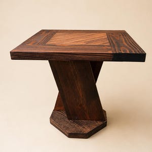 Puede incluir: Una mesa auxiliar cuadrada de madera con un acabado teñido en marrón oscuro. El tablero presenta un panel central con patrones de veta de madera en diagonal. La base consta de una columna de soporte inclinada sobre una placa hexagonal.