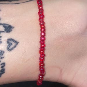 May include: A single-strand bracelet composed of small, round, translucent red glass beads. The beads are uniform in size and shape, strung together in a continuous line to form a simple, minimalist piece of jewelry worn around the wrist.