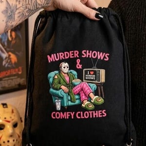 May include: A black drawstring backpack featuring a graphic of a hockey-masked figure in a green robe and pink plaid pants sitting in a blue armchair, eating popcorn, with the text "MURDER SHOWS & COMFY CLOTHES" and a television displaying "I LOVE HORROR SHOWS".
