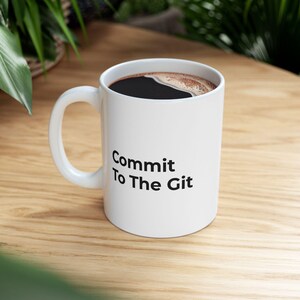 May include: White ceramic mug with the text "Commit To The Git" in black. The mug is filled with dark coffee. The mug is on a wooden surface.