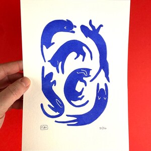 May include: A print featuring stylized blue cat illustrations in various poses on a white background. The artwork includes a signature and the number 21/50. The print is held against a red background.