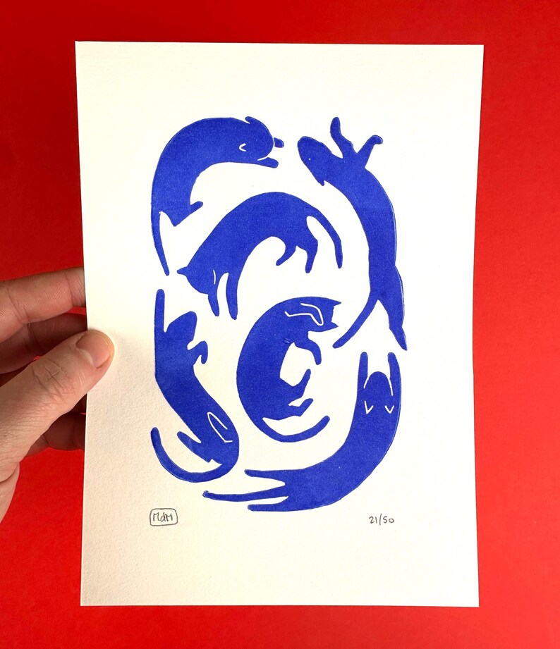 May include: A print featuring stylized blue cat illustrations in various poses on a white background. The artwork includes a signature and the number 21/50. The print is held against a red background.