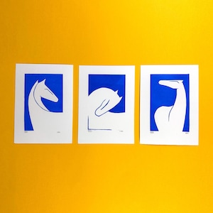 May include: Three white horse silhouette art prints with blue backgrounds, set against a yellow backdrop. Each print shows a different pose of a horse's head and neck. The prints are rectangular and have a minimalist design.