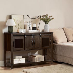 May include: A dark brown wooden console table with sliding doors and a lower shelf. The table has a rectangular shape and features black hardware. Decorative items, including a lamp, framed art, and a vase with flowers, are displayed on top.