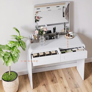Op de afbeelding: Een witte kaptafel met een rechthoekige spiegel, twee lades en een gebogen ontwerp. De lades zijn open en onthullen make-upproducten. Een potplant met groene bladeren staat links. De tafel staat tegen een witte muur.