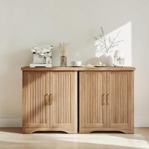May include: Two light-colored wooden cabinets with vertical grooved door panels and brass handles. A white espresso machine and a white appliance sit on top, along with a vase of dried flowers and a cup and saucer. The cabinets are against a white wall.