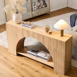 May include: A light brown wooden console table with an arched base. The table has a lower shelf and is decorated with a vase of flowers, books, candles, and a small lamp. The table is 48 inches long, 16 inches wide, and 30 inches high.