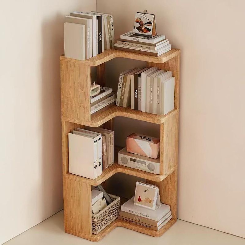 K&ouml;nnte beinhalten: Eckregal aus hellem Holz. Die Regale sind mit B&uuml;chern, Ordnern und Dekorationsartikeln gef&uuml;llt. Das Design ist eine moderne, platzsparende L&ouml;sung f&uuml;r die Organisation eines Home-Office oder Wohnraums.