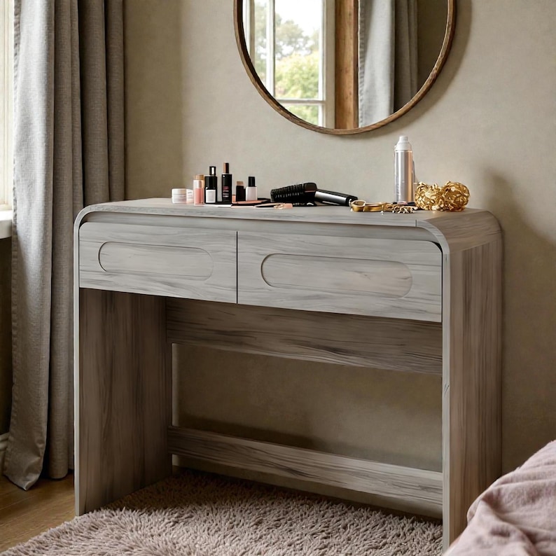 Op de afbeelding: Een lichtgrijze houten kaptafel met twee lades en een onderste plank. De tafel heeft een gebogen ontwerp en is bedekt met diverse schoonheidsproducten, een f&ouml;hn en decoratieve items. Een ronde spiegel hangt aan de muur boven de tafel.
