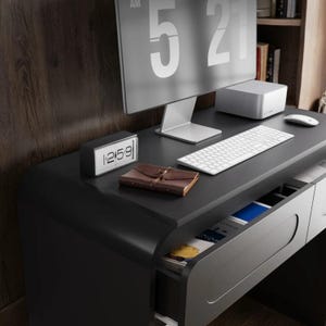 Op de afbeelding: Een moderne zwarte bureau met een computermonitor die 5:21 uur weergeeft. De bureau heeft een wit toetsenbord, muis en een kleine digitale klok met de tijd 12:59. Een bruin leren notitieboek ligt op de bureau, met een lade gedeeltelijk open.