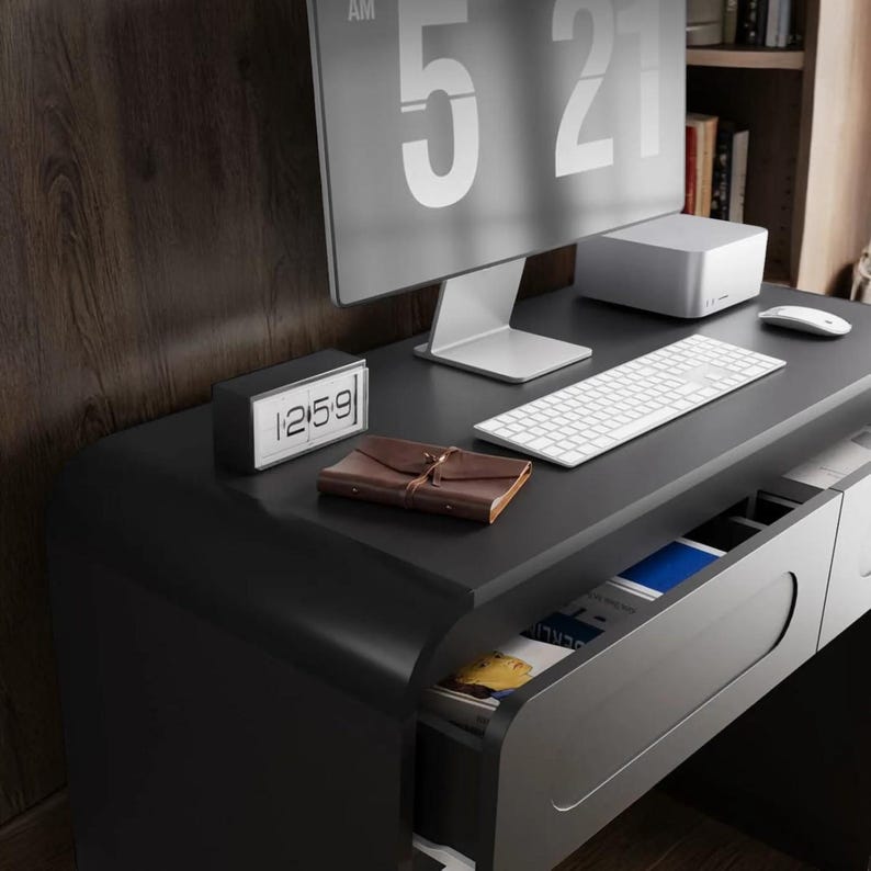 Op de afbeelding: Een moderne zwarte bureau met een computermonitor die 5:21 uur weergeeft. De bureau heeft een wit toetsenbord, muis en een kleine digitale klok met de tijd 12:59. Een bruin leren notitieboek ligt op de bureau, met een lade gedeeltelijk open.