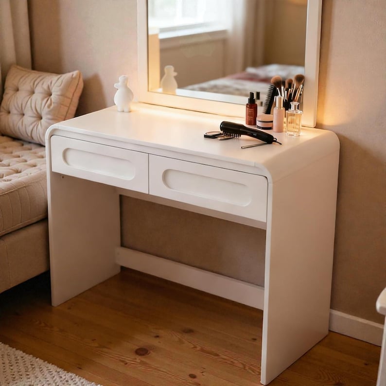 Op de afbeelding: Een witte kaptafel met twee lades en een gebogen ontwerp. Een spiegel is aan de bovenkant bevestigd en weerspiegelt een slaapkamersc&egrave;ne. Make-up borstels, flesjes en een haarborstel liggen op het tafelblad. Een klein wit beeldje staat links.
