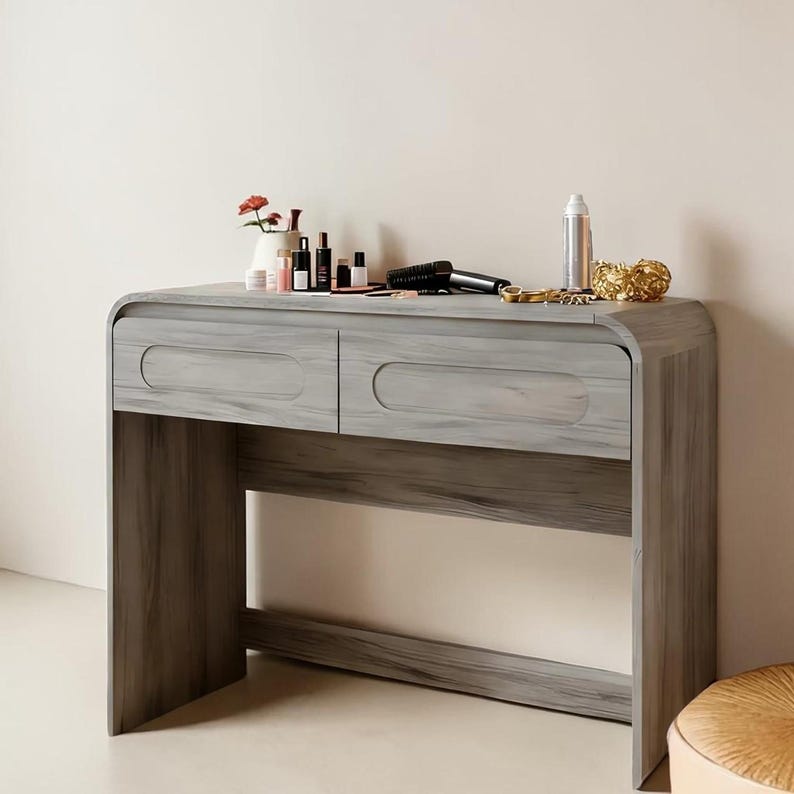 Op de afbeelding: Een grijze houten kaptafel met twee lades en een onderste plank. Het gebogen ontwerp heeft een licht houtnerfpatroon. Diverse schoonheidsproducten, een f&ouml;hn en decoratieve items zijn bovenop geplaatst.