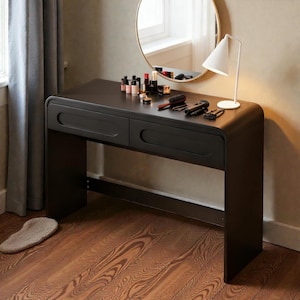 Op de afbeelding: Een zwarte kaptafel met twee lades, een ronde spiegel en een witte lamp. Make-up producten en borstels zijn op de tafel geplaatst. De tafel staat tegen een muur met een raam en een grijze gordijn.