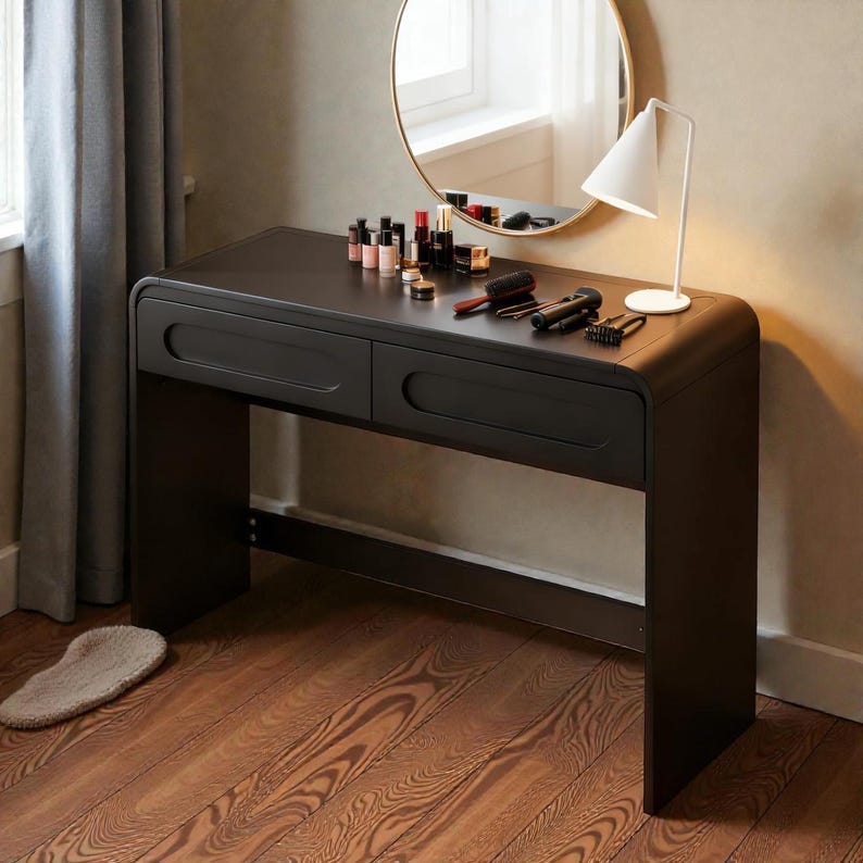 Op de afbeelding: Een zwarte kaptafel met twee lades, een ronde spiegel en een witte lamp. Make-up producten en borstels zijn op de tafel geplaatst. De tafel staat tegen een muur met een raam en een grijze gordijn.