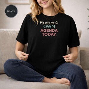 May include: A black short-sleeved t-shirt featuring the text "My body has its OWN AGENDA TODAY" printed in a mix of cream, teal, and pink colors. The shirt has a crew neckline and a relaxed fit, showcasing a clear graphic design on the front.