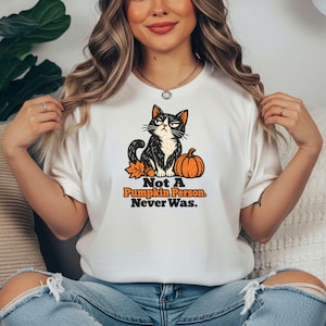 May include: A white short-sleeve t-shirt featuring a graphic of a black and white cat with a grumpy expression next to an orange pumpkin and an autumn leaf. The text below the cat reads, "Not A Pumpkin Person. Never Was."