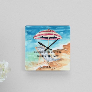 May include: Square wall clock featuring a beach scene with a red and white striped umbrella, a beach chair, and the text "Blessed is the one who trusts in the Lord Jeremiah 17:7". The clock has black hands.