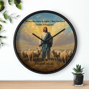 Op de afbeelding: Ronde wandklok met een zwart frame, met een religieuze scène van Jezus en schapen onder een bewolkte hemel. De wijzerplaat van de klok bevat de tekst "When the time is right, I the Lord will make it happen." en "Isaiah 60:22."