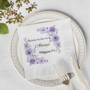 Può includere: Tovagliolo di carta bianco con la scritta "Rejoice in the Lord Always Philippians 4:4" e un motivo floreale viola. Il tovagliolo è posto su un piatto decorativo, con forchetta e coltello.