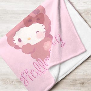 Puede incluir: Una manta rosa con un personaje de dibujos animados, un lazo y detalles florales. La palabra "Hello" está escrita en cursiva. La manta está doblada, revelando un reverso blanco.