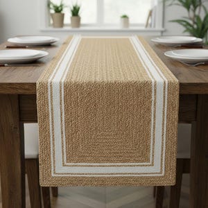 Op de afbeelding: Een rechthoekige tafelloper met een geweven textuur in een natuurlijke bruine kleur. Het heeft een witte rand en twee witte strepen in de lengterichting. De loper ligt op een houten tafel.
