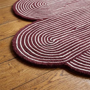 Pode incluir: Um tapete com padr&atilde;o bord&ocirc; e branco com uma forma &uacute;nica de nuvem. O tapete apresenta uma s&eacute;rie de linhas curvas conc&ecirc;ntricas em branco sobre um fundo bord&ocirc;. O tapete &eacute; feito de um material macio e texturizado.