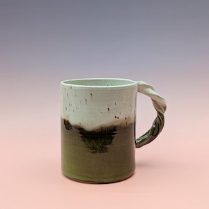 Peut inclure: Une tasse en céramique faite à la main avec une anse torsadée. La tasse présente un design bicolore, avec une partie supérieure blanche et une partie inférieure vert foncé. La anse est torsadée, ajoutant une touche unique à l'esthétique de la tasse.