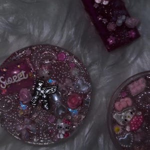 May include: A purple lighter and two pink resin coasters decorated with glitter, beads, and Hello Kitty figures. One coaster has the word "Sweet" on it. The items are on a white, fluffy surface.