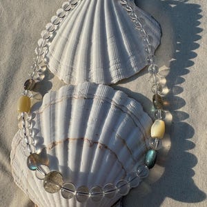 Puede incluir: Un collar de cuentas transparentes con una variedad de cuentas de piedras preciosas, incluyendo piedras transparentes, amarillas e iridiscentes. El collar se muestra sobre una concha blanca contra un fondo neutro. El collar se asegura con un cierre plateado.
