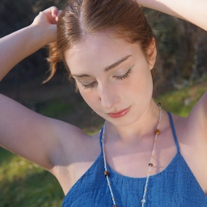 Può includere: Una donna indossa un top blu e una collana di perline con un ciondolo. La collana presenta perline trasparenti, marroni e dorate. La donna ha i capelli rossi e guarda in basso.