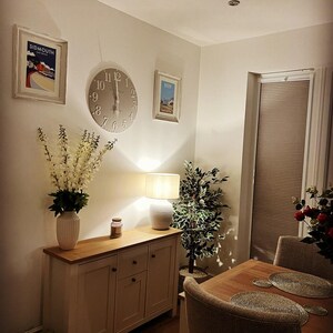 May include: A well-lit dining room with a wooden table, chairs, and a sideboard. The room features framed artwork, a large clock, a lamp, and a vase of flowers. A window with blinds is visible.