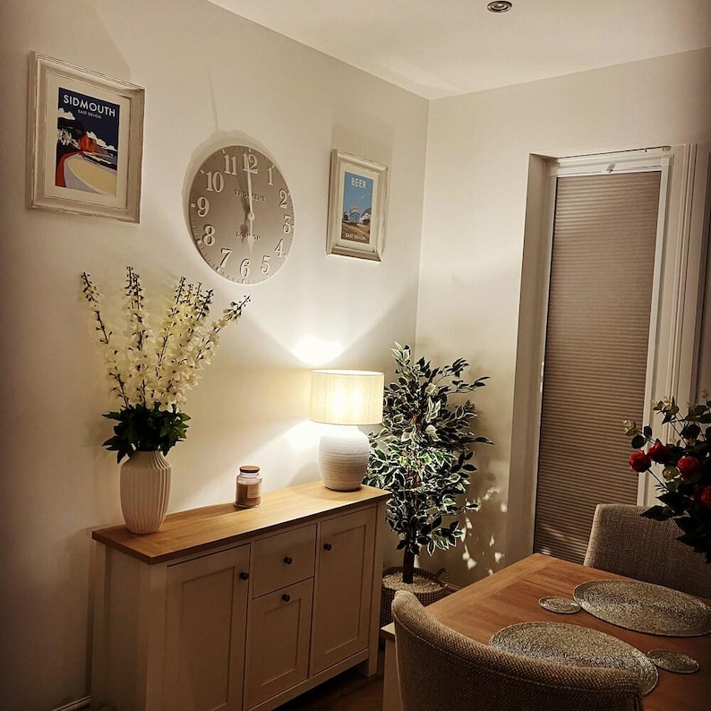 May include: A well-lit dining room with a wooden table, chairs, and a sideboard. The room features framed artwork, a large clock, a lamp, and a vase of flowers. A window with blinds is visible.