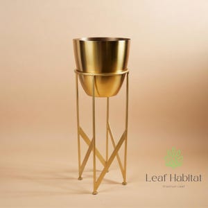May include: A gold metal planter on a tall stand with a geometric design. The planter has a rounded bowl shape, and the stand has a cross-braced design. The Leaf Habitat logo is visible in the bottom right corner.