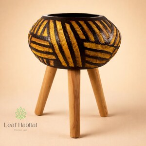 May include: A handcrafted wooden planter with a black interior and a geometric pattern in yellow and black. The planter is supported by three light-coloured wooden legs. The logo "Leaf Habitat" is visible.