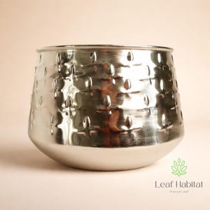 Puede incluir: Una maceta de metal plateado con textura martillada y base redondeada. La maceta tiene una abertura ancha y un patrón de pequeñas hendiduras alargadas. El logotipo de Leaf Habitat es visible en la esquina inferior derecha.