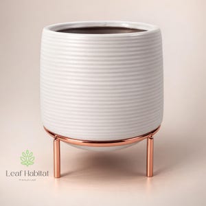 May include: White ceramic plant pot with a ribbed design, supported by a copper-toned metal stand. The pot has a cylindrical shape and a dark interior. The stand features a circular base and three vertical legs.
