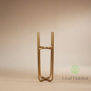 May include: A gold metal plant stand with a contemporary design. The stand has a cross base, vertical supports, and a bow-shaped accent. The Leaf Habitat logo is in the lower right corner.