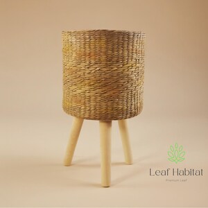 May include: A cylindrical woven basket planter, supported by three pale wooden legs. The basket is made from natural-coloured woven material. The Leaf Habitat logo is in the lower right corner.