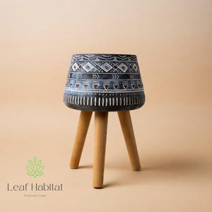 May include: A small, decorative plant stand with a dark blue, patterned top and three wooden legs. The top features a geometric design in white and brown. The stand is approximately 30 cm tall.