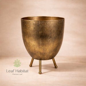 May include: A gold-coloured metal planter with a textured finish. The planter has a rounded, egg-shaped body and stands on three slender legs with rounded feet. The planter is designed for indoor or outdoor use.