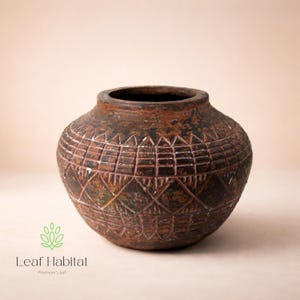 May include: A weathered, brown ceramic pot with a wide opening and a rounded body. The pot has a detailed geometric pattern carved into its surface. The pot has a rustic appearance, suggesting age and use. The Leaf Habitat logo is at the bottom.