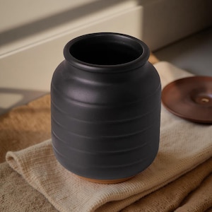 Puede incluir: Un jarrón de cerámica negro con detalles horizontales acanalados. El jarrón tiene una abertura ancha y se asienta sobre una base marrón. Una pequeña tapa redonda marrón está al lado del jarrón. El jarrón está sobre una tela beige.