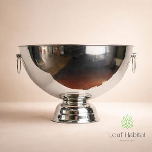 May include: A large, polished silver metal bowl with two ring handles. The bowl sits on a pedestal base. The bowl reflects the surrounding environment. The Leaf Habitat logo is in the bottom right corner.
