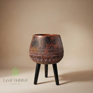 May include: A decorative, brown and black planter with a tribal pattern, supported by three black legs. The planter has a textured surface and a wide opening. The Leaf Habitat logo is visible in the lower left corner.