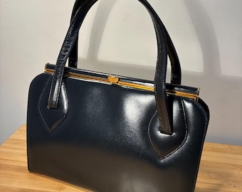 Puede incluir: Un bolso de mano vintage azul marino con una silueta estructurada. El bolso presenta dos asas arqueadas, un cierre dorado y un acabado brillante. El bolso está sobre una superficie de madera.