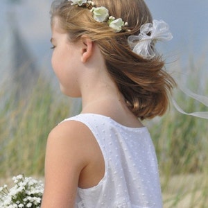 May include: A young girl wearing a white dress with a floral crown and a white bow in her hair. She is holding a bouquet of white flowers.